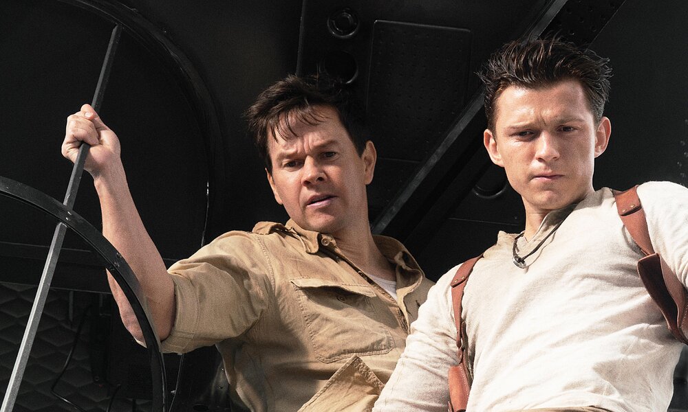 Nahaufnahme von Mark Wahlberg und Tom Holland als Schatzsucher in einer Szene aus dem Kinofilm "Uncharted". Sie stehen vor dunklem Hintergrund nah nebeneinander und blicken konzentriert nach unten. | © ddp images/PictureLux
