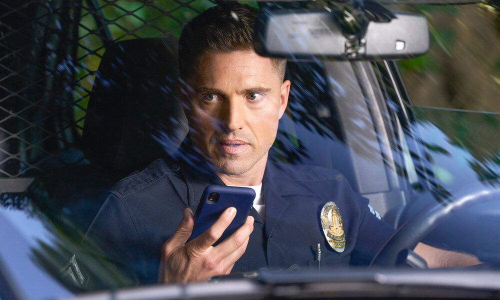Blick durch die Windschutzscheibe eines Polizeifahrzeugs auf Eric Winter als Police Officer Tim Bradford in dunkelblauer Uniform mit goldenem Abzeichen, der in ein Smartphone spricht in einer Szene aus der ABC-Serie "The Rookie" | © Getty Images/Scott Everett White 