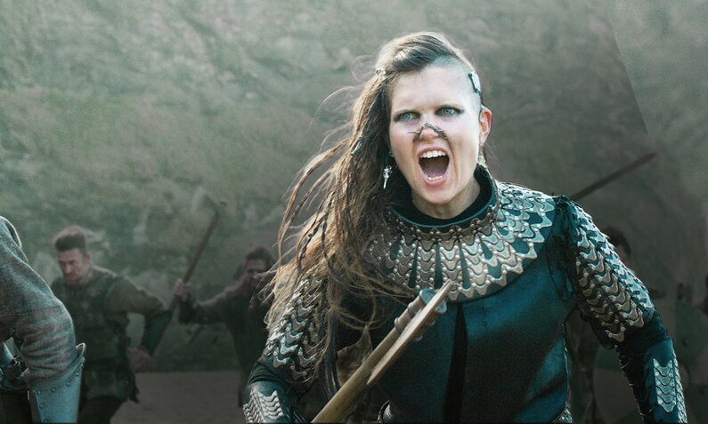 Junge Frau mit wehendem langen Haar in altertümlicher Rüstung mit gezogenem Schwert stürmt in einer Szene der Serie "Cursed" zusammen mit Männern. im Hintergrund ein Berg. | © Netflix