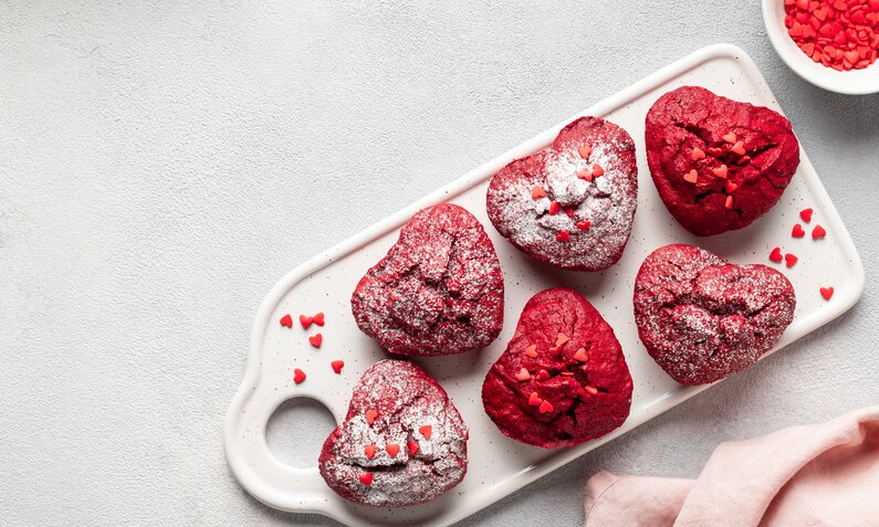 Rote Muffins in Herzform, bestreut mit Streuseln auf weißen Untergrund. | © Getty Images / Mila Naumova