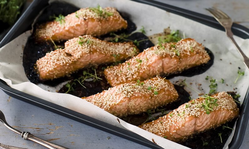 Lachsfilets mit Sesamkruste auf einem Backblech angerichtet, dahinter frischer Basilikum. | © Getty Images / Tasty food and photography