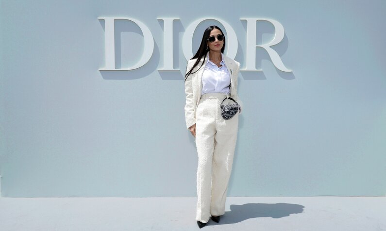 Demi Moore steht in weißen eleganten Anzug vor einer grauen Wand mit dem weißen Schriftzug "Dior" vor der Männermodenschau 2024 in Paris. | © Getty Images/Pascal Le Segretain