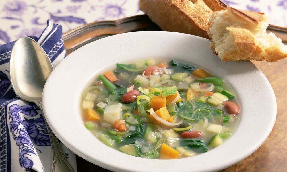 Gemüsesuppe angerichtet in weißem Suppenteller, daneben Baguette-Scheiben und ein Löffel. | © Getty Images / 	Brian Hagiwara