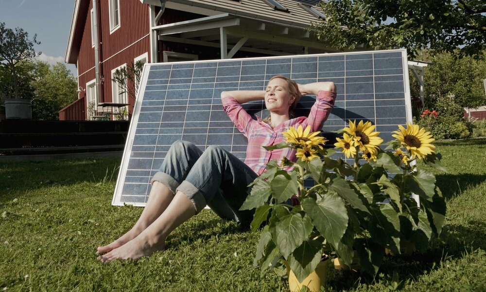 Frau sitzt glücklich in ihrem Garten und lehnt sich an ein Solarpanel an | © Getty Images/Robert Niedring