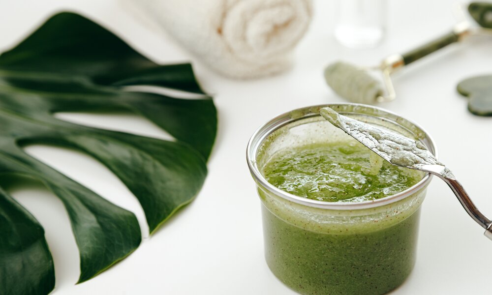 Grüne Creme in Glastiegel, daneben Monstera-Blatt. | © Getty Images / 	Vladdeep
