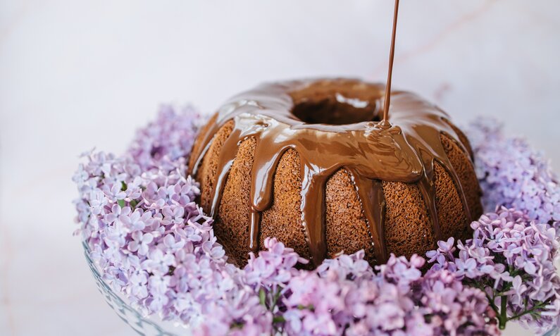 Schoko-Gugelhupf-Kuchen, begossen mit Schokoladensoße, angerichtet auf einem Blütenbett. | © Getty Images / 	Vuk Saric