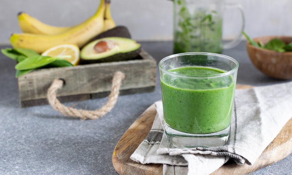 Grüner Smoothie in Glas, dahinter Bananen, Avocado und Zitrone liegend in Holzkorb. | © Getty Images / Anastasia Turshina