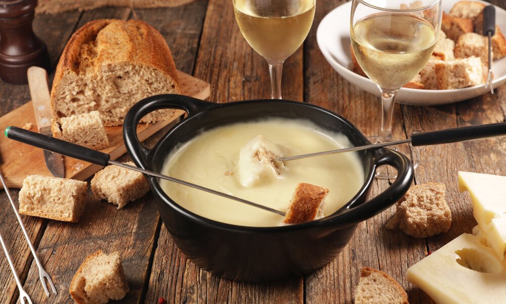 Ein Fondue-Set mit geschmolzenem Käse darin, Hände halten Lebensmittel an Stäben in den Käse, daneben Weißwein und Brot. | © Getty Images / 	margouillatphotos
