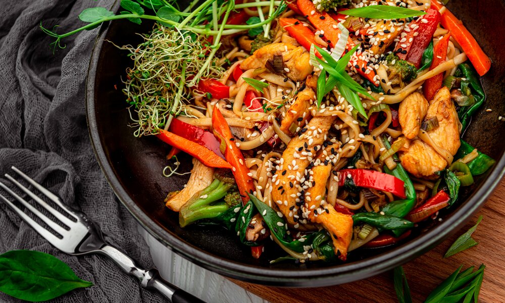 Putengeschnetzeltes mit Paprika, Sesam, Frühlingszwiebeln und anderen Zutaten, angerichtet in einem Wok, daneben Besteck. | © Getty Images / YuraWhite
