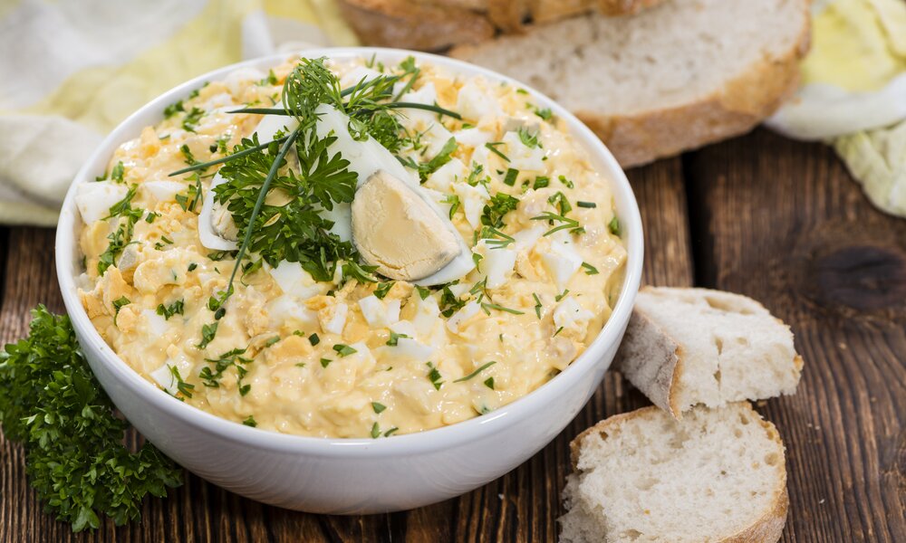 Eiersalat in weißer Schüssel, bestreut mit frischer Petersilie, daneben Brotscheiben. | © Getty Images / 	HandmadePictures