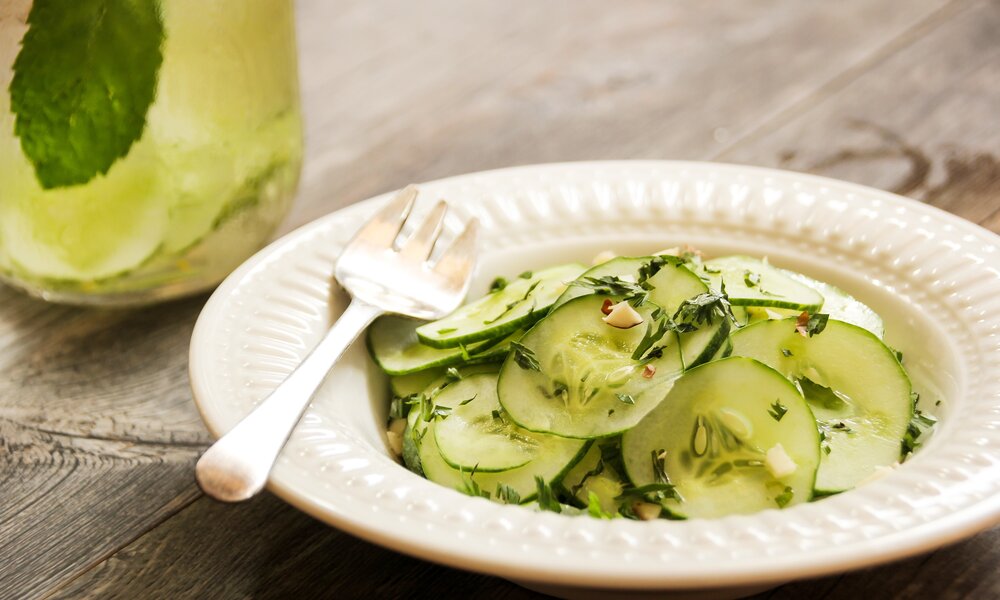 Gurkensalat mit Dill, angerichtet auf weißem Teller, daneben Gabel und ein Glas mit einem gelblichen Getränk. | © Getty Images / 	vm2002