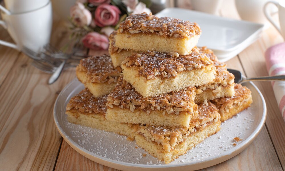 Streuselkuchenstücke drapiert auf einem weißen Teller auf einem Tisch, dahinter Blumen. | © Getty Images / 	Angelika Heine