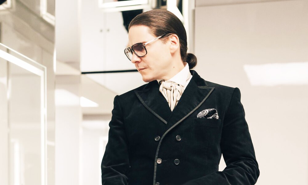 Daniel Brühl steht als Modedesigner Karl Lagerfeld in eleganter Kleidung in einem hell beleuchteten, hellen Raum und blickt zur Seite in einer Szene aus der Disney+ Serie "Becoming Karl Lagerfeld". | © Disney+