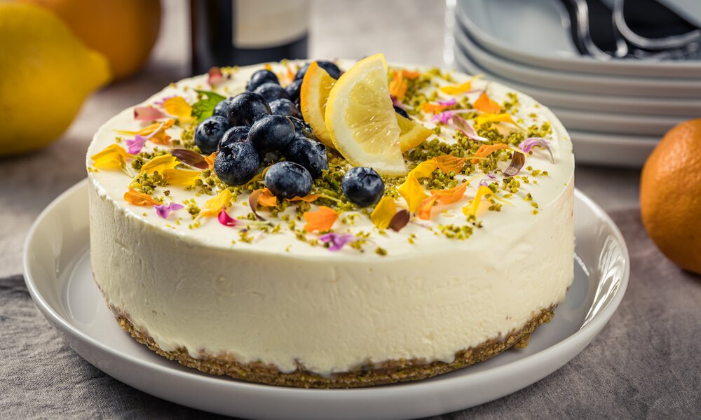Frischkäse-Torte mit Keksboden auf einem Holzuntergrund, dekoriert mit vielen bunten Frühlingsblüten, Blaubeeren und einer Zitronenscheibe. | © Getty Images / 	Pixsicle Photography