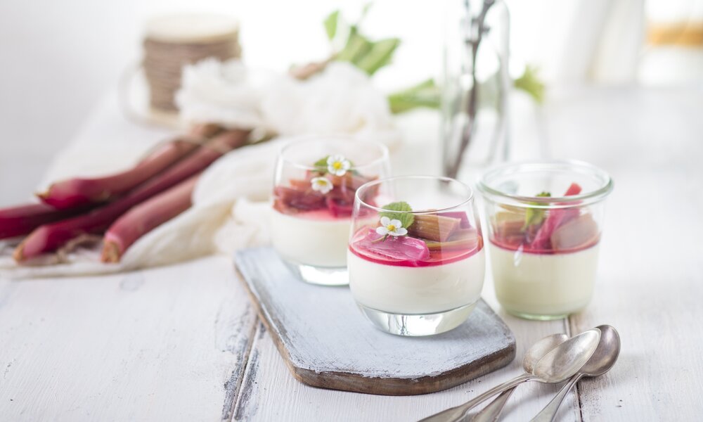 Weiße Creme geschichtet in Dessertglas, on top Rhabarberkompott. Frühlingshafte Atmosphäre und Blüten im Hintergrund. | © Getty Images / 	Westend61