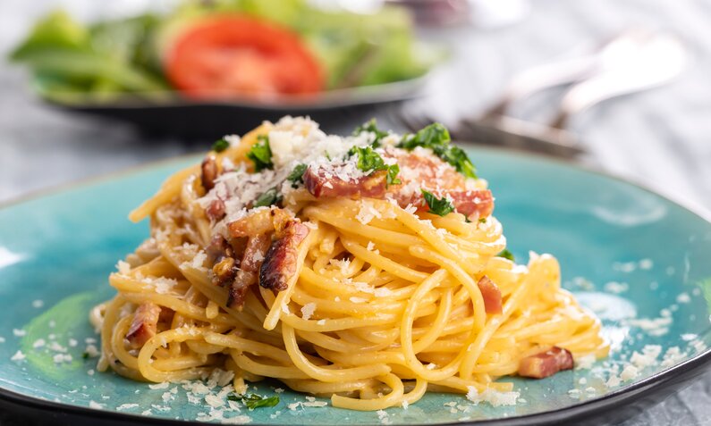Aufgedrehte Spaghetti Carbonara auf einem Teller, darüber gestreut knuspriger Speck. | © Adobe Stock / Bernd Jürgens