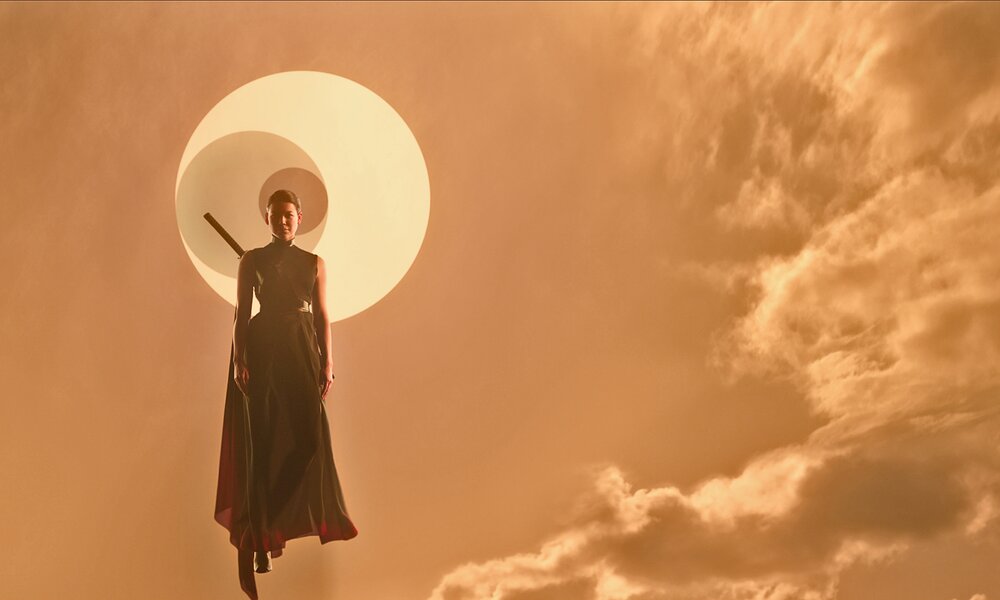 Sea Shimooka schwebt als Sophon vor einem orangeroten Himmel mit Wolken und bleichem Mond in einer Szene aus der Netflix-Serie "3 Body Problem" | © Netflix