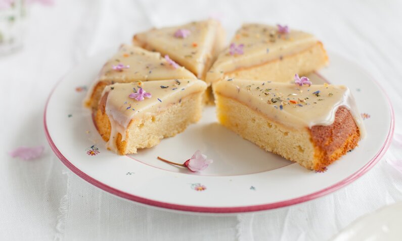 Zitronenkuchen in Stücke geschnitten, auf einem weißem Teller, bestreut mit bunten Blütenblättern. | © Getty Images / ingwervanille