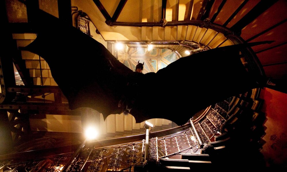 Christopher Bale ist in einer Szene aus dem Kinofilm "Batman Begins" zu sehen, wie er im Batmankostüm mit ausgebreitetem Cape einen Treppenschacht hinunterstürzt. | © Imago Images/United Archives