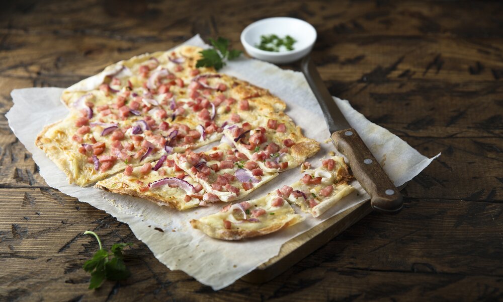 Flammkuchen in Stücke geschnitten auf einem Holzbrett, danebenliegend ein Messer und frische Petersilie. | © Getty Images / 	Mariha-kitchen