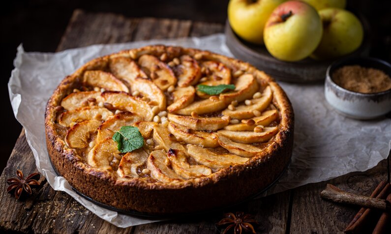 Apfelkuchen auf Holzuntergrund, daneben frischer Apfel und etwas frische Minze. | © Adobe Stock / petrrgoskov