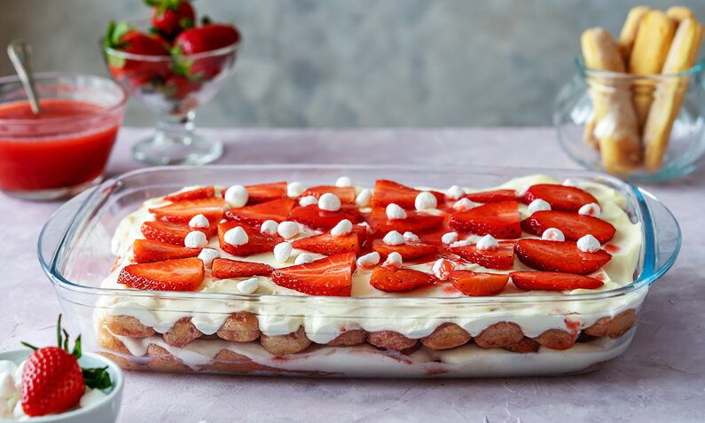 Erdbeer-Tiramisu, hergestellt im Thermomix in einer Glasform auf einem Tisch stehend. Zu sehen sind außerdem die enthaltenen Löffelbiskuits, frische Erdbeeren und Erdbeersoße. | © Getty Images / Tetiana Zavgorodnia