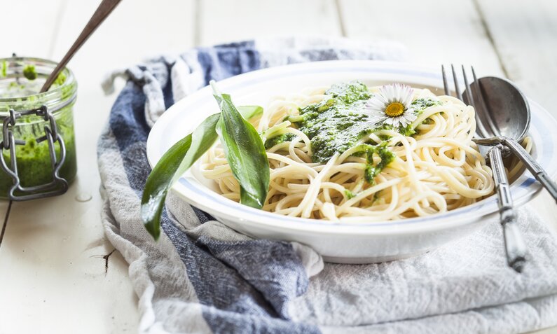 Grünes Bärlauch-Pesto auf einem Haufen Spaghetti auf einem weißen Teller, garniert mit frischen Bärlauchblättern, auf einem weiß-blauen Geschirrtuch stehend. Daneben steht ein Glasbehälter mit Pesto und eine Gabel und ein Messer. | © Getty Images / 	Westend61