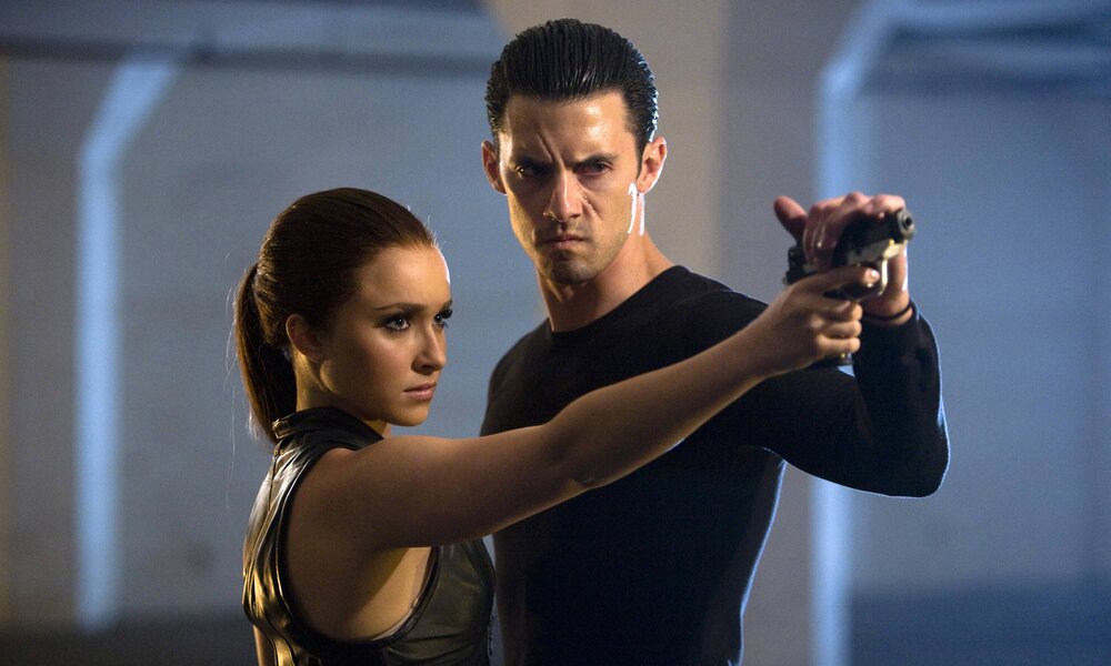 Hayden Panettiere und Milo Ventimiglia stehen mit ernsten Gesichtern in einer Szene aus der TV-Serie "Heroes" nebeneinander, sie hat eine Waffe. | © Imago Images/Everett Collection
