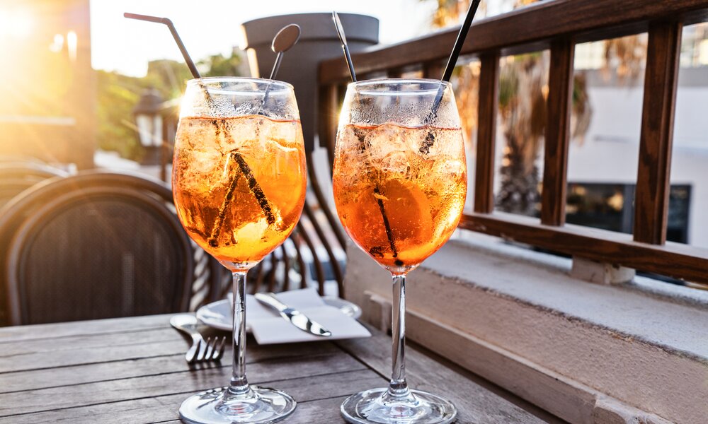 Zwei Gläser befüllt mit Aperol Spritz auf einem hölzerner Balkontisch stehend, mit je einem schwarzen Strohhalm innen. Leichte Sonneneinstrahlung von links. | © Adobe Stock / Kira_Yan