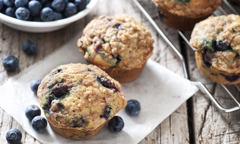 Mehrere Blaubeer-Muffins auf einem Backpapier und einem Gitterrost daneben. Außerdem sind frische Blaubeeren in einer weißen Schale oder lose auf dem Holzuntergrund zu sehen. | © Getty Images / 	mphillips007