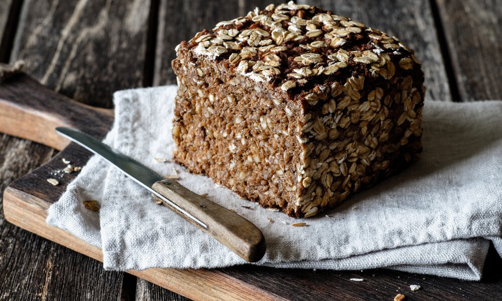 Aufgeschnittenes Vollkornbrot ohne Mehl und ohne Hefe auf einem weißen Geschirrtuch. Das Tuch liegt auf einem Holzuntergrund und daneben liegt ein Brotmesser. | © Adobe Stock: ji_images
