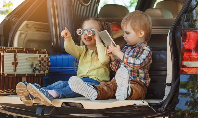 Lachendes Mädchen mit Fernglas-Spielzeug und Junge mit Tablet im Kofferraum eines Autos. | © Friends Stock /stock.adobe.com