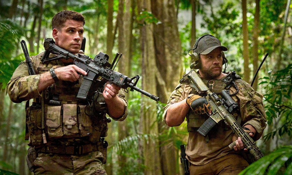 Liam und Luke Hemsworth schleichen in Tarnkleidung mit Gewehr im Anschlag durch den Wald. | © Imago Images/Landmark Media