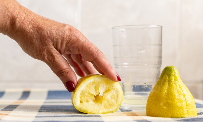 Halbe Zitrone wird auf blau-weiß kariertem Geschirrtuch ausgepresst, im Hintergrund eine ganze Zitrone und Wasserglas. | © Getty Images/ David Petrus Ibars