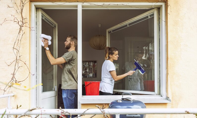 Pärchen bei Hausarbeit, sie zieht Fensterabzieher, er wischt Fenster von außen. | © Getty Images/ Maskot