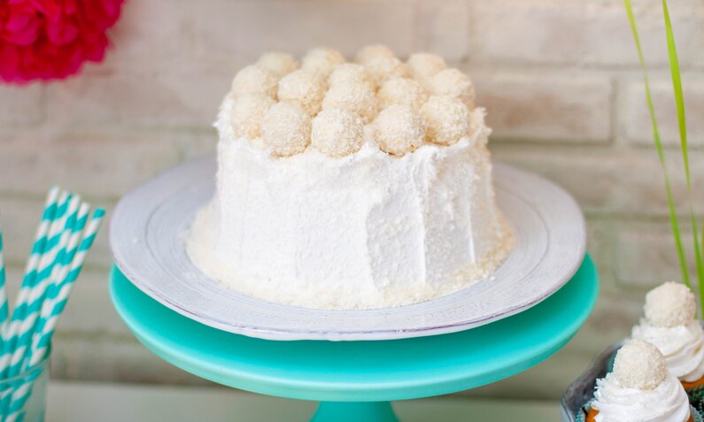 Weiße Raffaello-Torte ohne Backen auf einer weißen Kuchenplatte angerichtet vor weißem Hintergrund und garniert mit einigen Raffaello-Pralinen. | © Getty Images / 	Larisa Sadomtseva