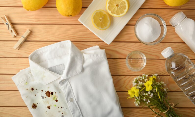 Weißes Hemd mit Flecken neben Zitronen und Hausmitteln auf Holztisch. | © Getty Images/ Davizro