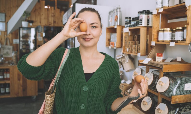 Frau mit brauen Haaren und grünem Pullover steht in einem Laden und hält sich ein braunen Ei vor ein Auge, um zu erkennen ob das Ei roh oder gekocht ist. | © Getty Images / Westend61