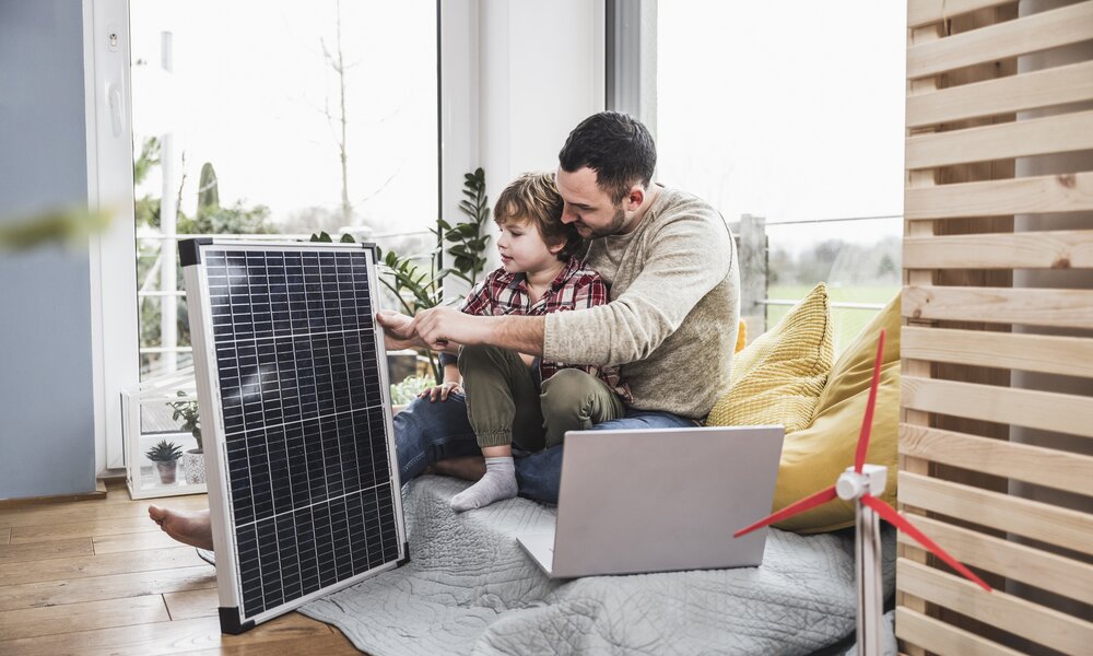 Vater und Sohn betrachten lernend ein Solarmodul neben einem Laptop im Wohnzimmer. | © Getty Images/Westend61