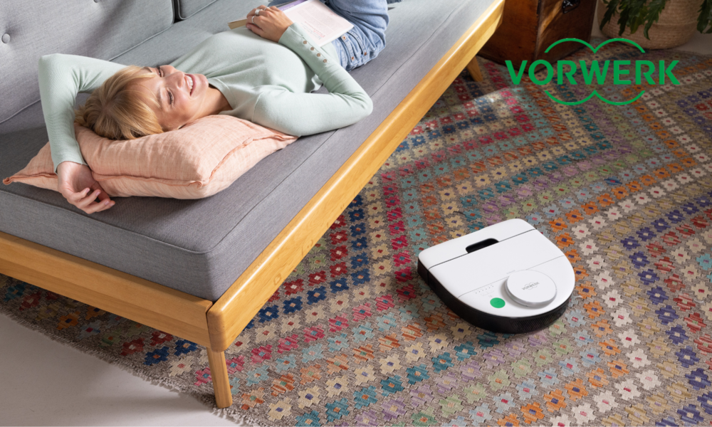 Lächelnde junge Frau liegt auf Couch, der Vorwerk Kobold RK7 Saugroboter arbeitet auf gemustertem Teppich im Wohnzimmer. | © Vorwerk