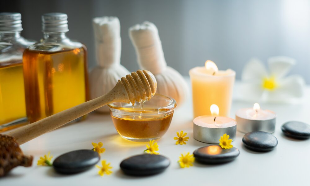 Honig fließt von Holzlöffel in Schale, umgeben von Massageölen, Kerzen und Blumen für Wellness. | © Getty Images/ Teera Konakan
