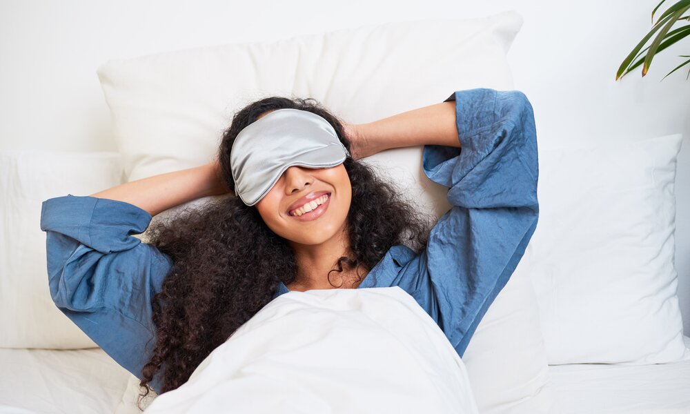 Eine Frau mit lockigen Haaren liegt lächelnd im Bett und hat eine graue Schlafmaske auf. | © Getty Images/Meeko Media