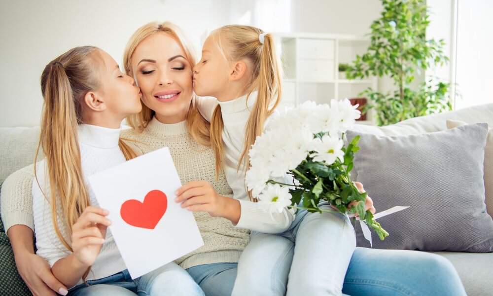 Fröhliche Mutter genießt Küsschen ihrer Töchter auf der Couch, hält rotes Herz und Blumenstrauß. | © deagreez/stock.adobe.com