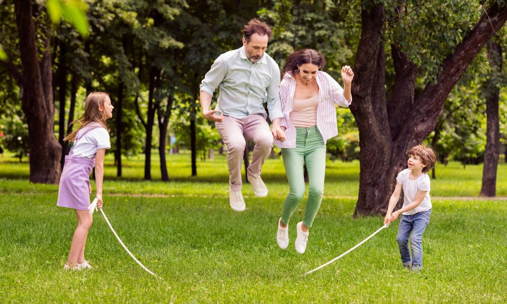 Familie spielt glücklich Seilspringen im Park, umgeben von saftigem Grün und alten Bäumen. | © deagreez /stock.adobe.com