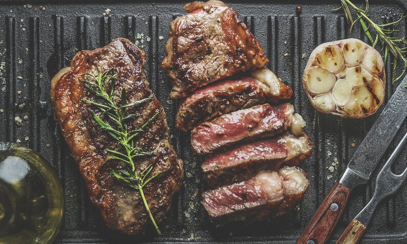 Zwei Steaks auf dem Grill, eines angeschnitten, das andere ganz, daneben gerösteter Knoblauch aufgeschnitten. | © Getty Images / VICUSCHKA