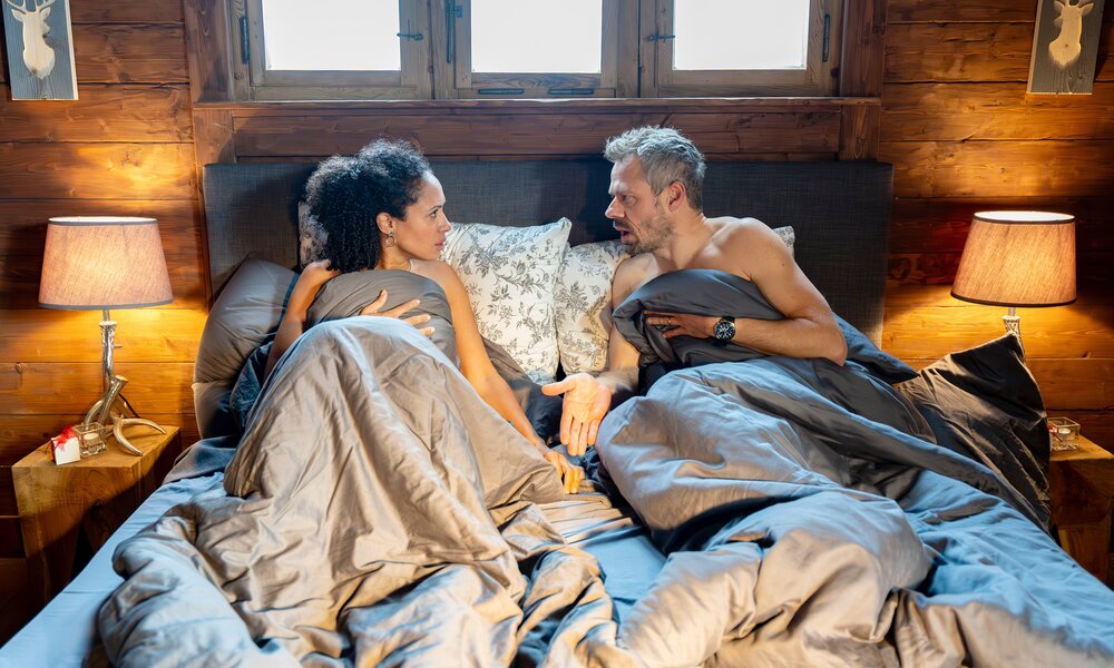 Eine lockige, dunkelhaarige Frau und ein grauhaariger Mann liegen im Bett unter einer grauen Bettdecke und schauen sich entsetzt an. | © ARD/WDR/Thomas Neumeier