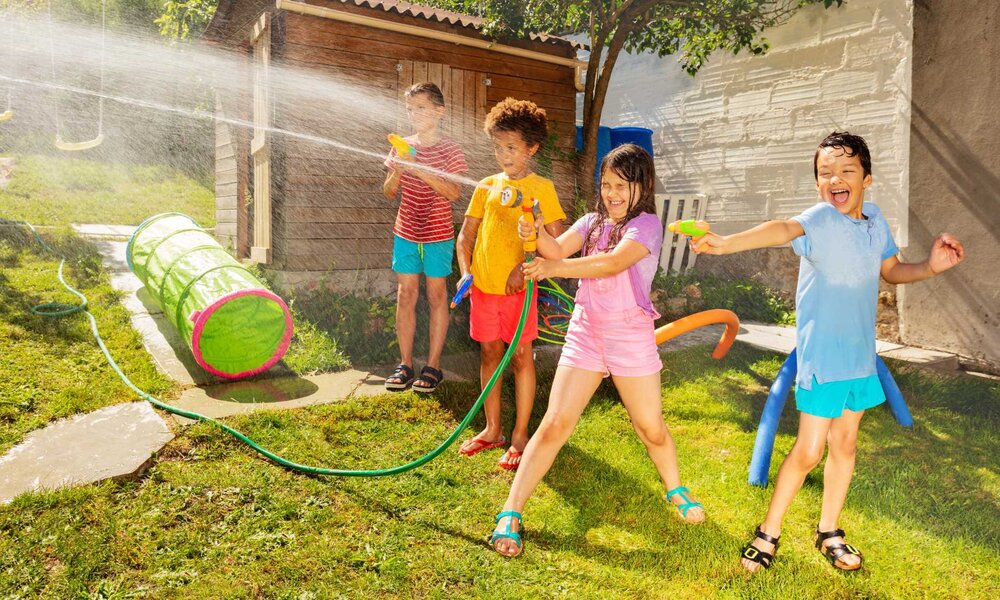 Vier Kinder spielen lachend mit Wasserpistolen im sonnigen Garten, grüne Wiese und hölzernes Spielhaus im Hintergrund. | © Sergey Novikov/stock.adobe.com