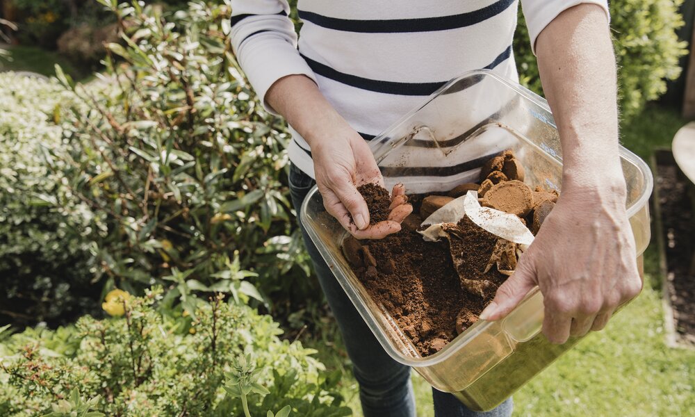 Frau steht mit einer Kiste Kaffeesatz vor einem Beet | © Getty Images/ DGLimages