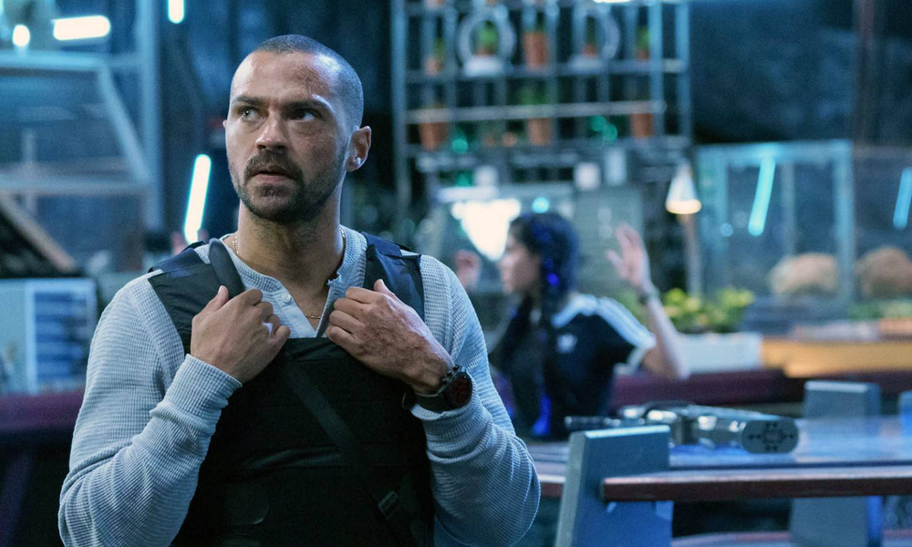 Jesse Williams steht mit schusssicherer Weste und ernstem Gesicht in einer blau beleuchteten Halle in einer Szene aus dem Film "Secret Headquarters" | © Imago Images/Everett Collection
