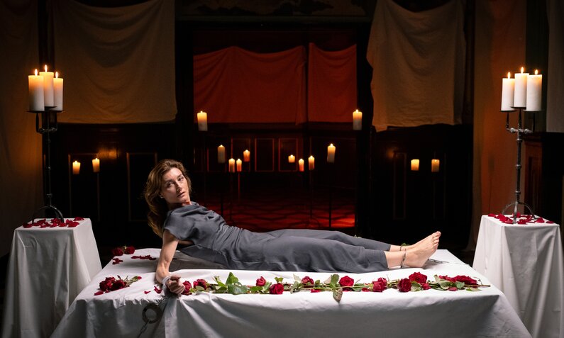 Brünette Frau in grauem Kleid liegt auf einem Tisch, umringt von Rosen und Kerzen in einem dunklen Raum. | © ARD/WDR/Christof Arnold